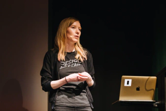 A closeup of a CSSConf speaker, Diana Mounter, holding her hands together while presenting. She has blonde hair past her shoulders, wearing a grey t-shirt with 'Design details' lettering and a cardigan. Her laptop is also visible on a lantern.