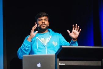 A closeup of a JSConf speaker, Vigneshwer Dhinakaran, holding both hands up while presenting. He's wearing a vibrant blue jumper and a t-shirt.