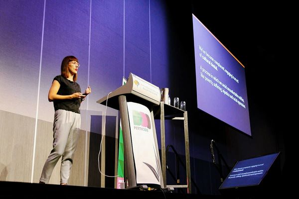 Karolina speaking at DDD in Perth. She's on a large stage adjacent to a lectern with a laptop. There's a small screen with her slides, acting as speaker notes, and a large screen displaying the slides to the audience. She's wearing grey pants and black t-shirt and is mid-talking.