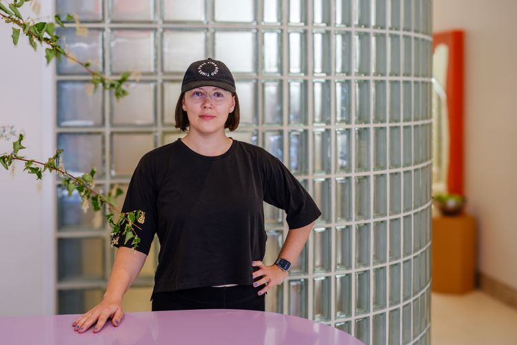 Portrait of Karolina standing with one hand on the hip, and one on a whimsical, blob-shaped purple desk in front of her. She’s wearing a black t-shirt, pants and a cap that reads 'Full Time Good Times'. She’s smiling and wearing optical glasses. Behind her, there’s a wall made of glass squares. She’s in her favourite pilates studio, Good Times Pilates.
