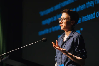 A closeup of a CSSConf speaker, Ivana, holding one hand up near the microphone as the presents. She's wearing a dark shirt, glasses, and has short hair.