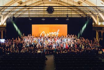 The main conference room, facing the stage and big display with a CSSConf logo. In front of the display, there's the majority of attendees, speakers and organisers smiling and waving towarsds the camera. There are probably 200 people there.