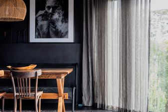 Interior of a cottage in Anglesea. Cropped photo of a dining room painted black with a timber table, a large black-and-white portrait, a rattan pendant, and sheer linen curtains opening onto bushland.
