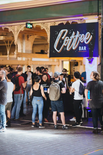 The entry room of the Meat Market with dozens of attendees in front of the coffee bar. The bar has a large, black sign with white lettering spelling 'Coffee bar' in a hand brush style.