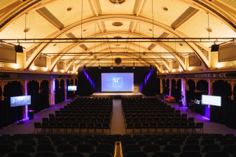 The main conference room while empty, facing the stage and big display with a JSConf logo. There's auditorium-like seats in the middle and TV displays for captioning visible to the sides.