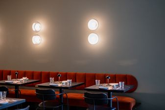 Interior of Kaya motel in Blue Mountains. Red velvet banquette seating along a soft grey wall, with set black tables and four glowing disc sconces.