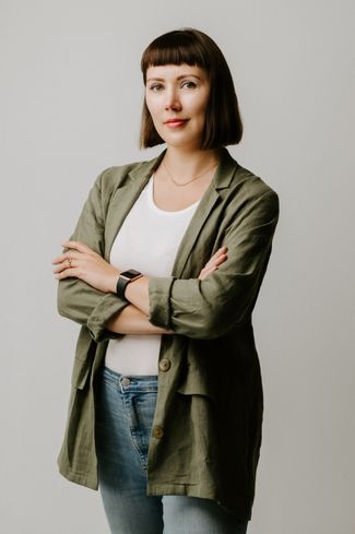 Portrait of Karolina on a grey background. She's wearing a khaki green jacket on a white t-shirt with jeans. She's slightly smiling with her arms friendlily crossed across her body.