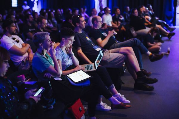 A dark room filled with CSSConf audience, focused on first rows of attendees. The main focus of the photo are two women, one listening attentively while holding her face, the other taking notes on her laptop.