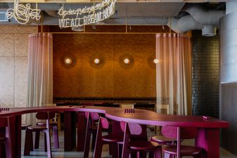 Interior of Lola Pizza in Kraków. Fit-out in crimson and gold: curved dark magenta counter and stools beneath a neon script sign saying 'We all dream about Lola' and an amber wall.