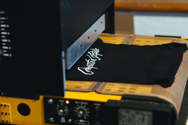 A closeup of screen printer showing a black women's cut t-shirt having 'Computed Style' printed on the chest area.