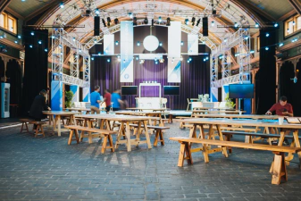 The entry room of the Meat Market. There is sponsor signage handing from a scaffolding by the ceiling. There's also cobblestone floor and wooden, beer-garden like tables waiting for attendees to flow in.