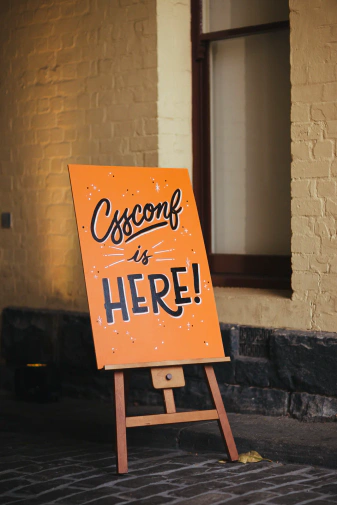 Trestle with an A-frame, vibrant orange sign with black hand lettering saying 'CSSConf is here!'